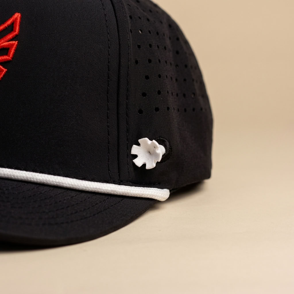 Close-up of a black baseball cap with a white rope detail along the brim, a small white flower-shaped embellishment, and a red embroidered logo on the side, set against a solid beige background.