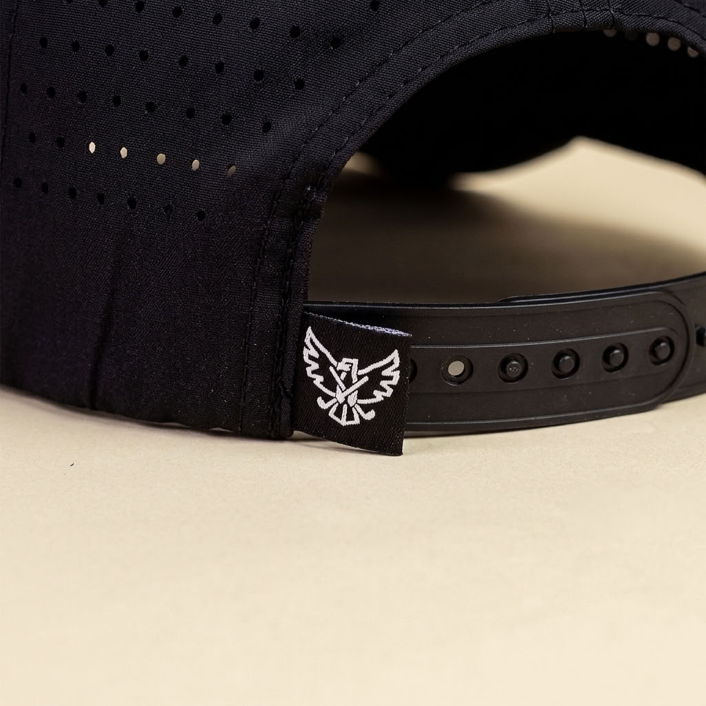 Close-up of the back of a black baseball cap with a black adjustable strap and a small black fabric tag featuring a white eagle logo, set against a light beige background.